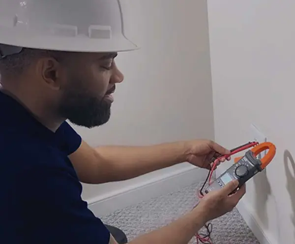 A close-up of a technician's hands using a digital multimeter to test a white electrical wall outlet.