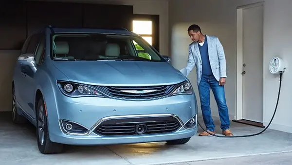 A man in a blue sweater and light gray blazer plugs a charging cable into a light-blue Chrysler Pacifica minivan parked inside a clean garage. The charging cable is connected to a white, wall-mounted electric vehicle charging station near a closed door.