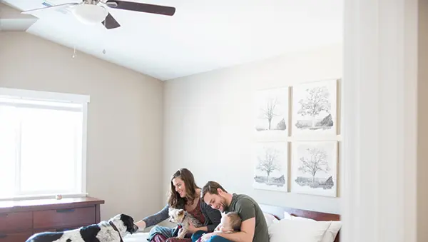 A family of three—a man, a woman, and a baby—sit together on a bed alongside two dogs. The room has neutral beige walls, four framed tree art prints, and a vaulted ceiling with a white-and-wood five-blade ceiling fan running above them.
