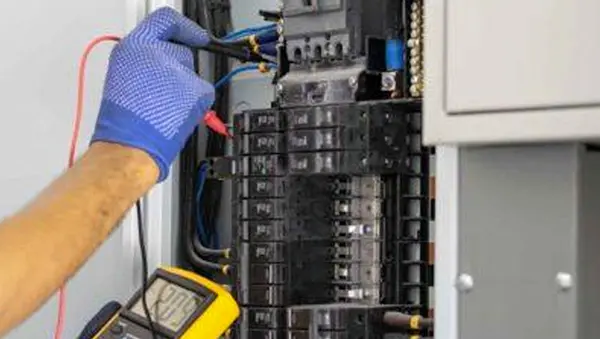Close-up of a person wearing a blue textured safety glove using a digital multimeter to test an open electrical circuit breaker panel. The yellow multimeter screen in the lower-left corner displays a reading of 120.9.