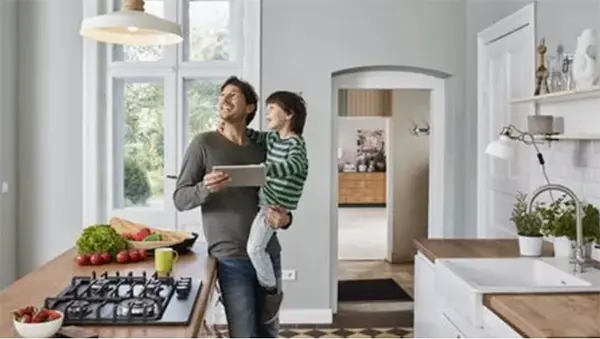 A smiling man standing in a modern kitchen holds a young boy in his arms while holding a digital tablet. Both look up and to the left toward a hanging light fixture. The kitchen features light gray walls, a wood countertop island with a built-in gas stovetop, and a white farmhouse-style sink.